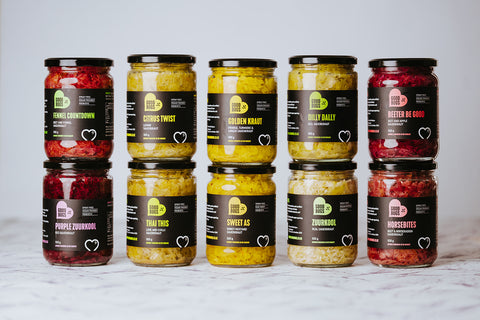 A selection of jars of GoodBugs sauerkraut with various labels, arranged in two rows on a light background. The sauerkraut jars come in different colors and are labeled with different flavors such as 'Golden Kraut', 'Zuurkool', 'Citrus Twist', and others.